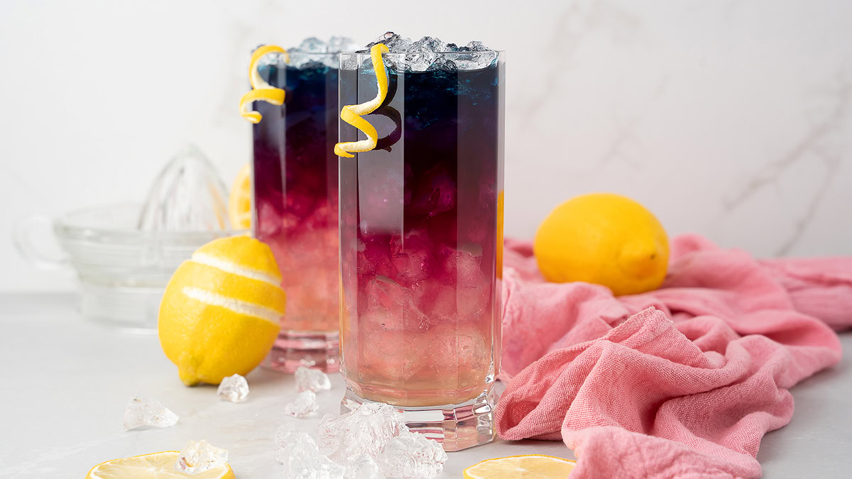 Two tall glasses filled with colorful layered drinks and ice, garnished with lemon twists, sit on a white surface with lemons, ice cubes, and a pink cloth nearby.