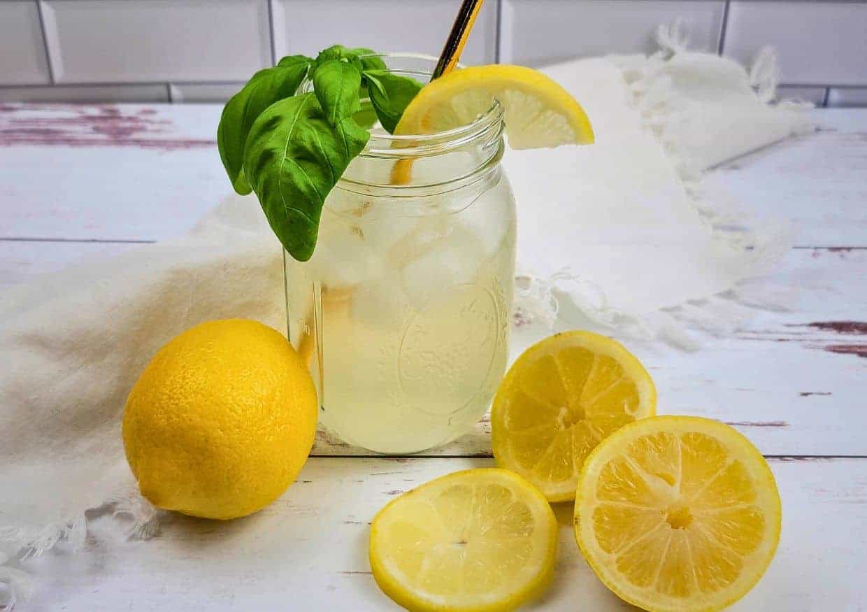 A mason jar of iced lemonade with a lemon wedge and basil garnish, surrounded by whole and sliced lemons on a white wooden surface.
