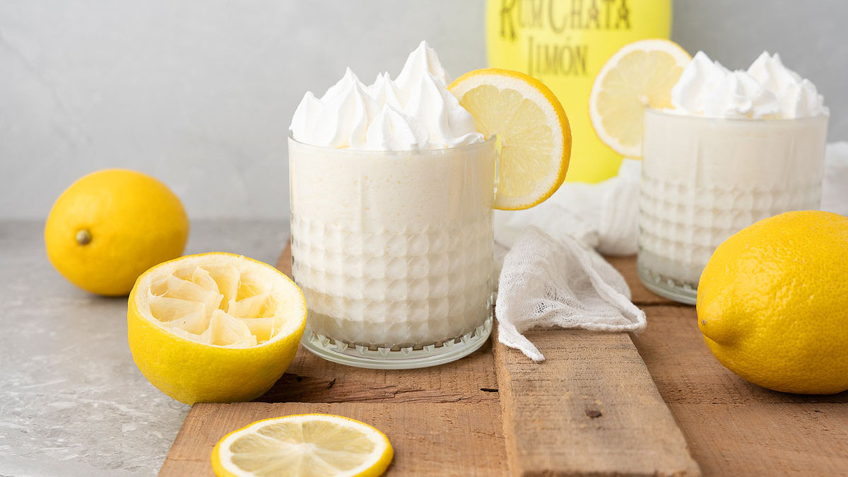 Two glasses of creamy white beverage topped with whipped cream and lemon slices, surrounded by whole and sliced lemons on a wooden surface.
