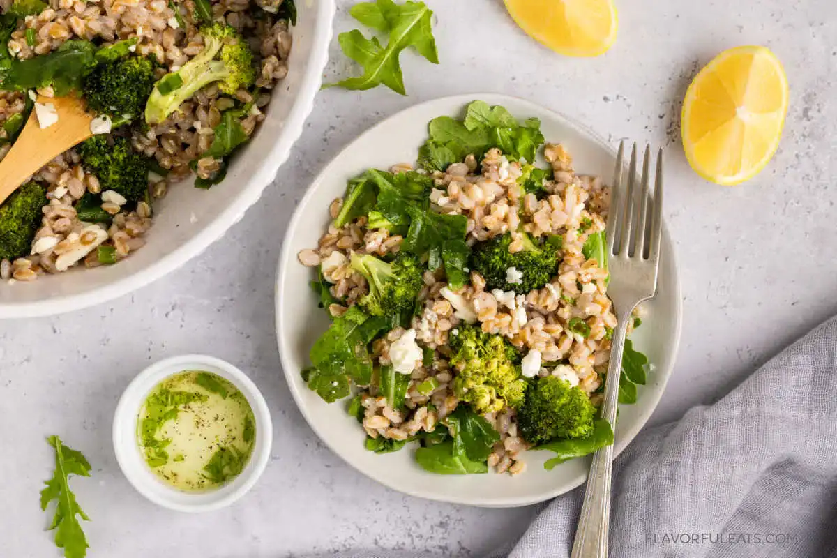 A plate of farro salad with broccoli, arugula, and crumbled cheese sits next to a small bowl of dressing, lemon wedges, and a gray napkin on a light surface.