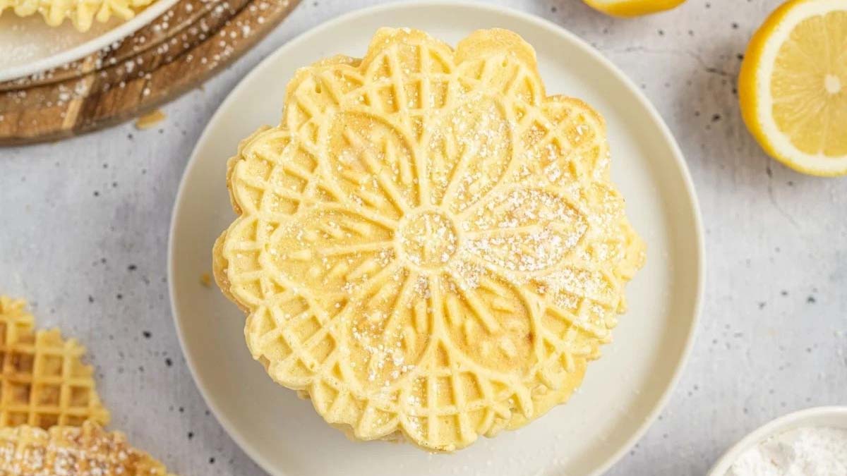 A stack of round, flower-patterned pizzelle cookies dusted with powdered sugar on a white plate, with lemon slices nearby.