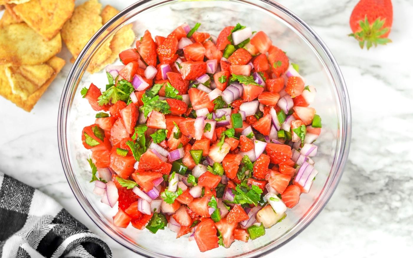 A glass bowl filled with strawberry salsa made of diced strawberries, red onion, cilantro, and jalapeño, placed on a marble surface with tortilla chips nearby.