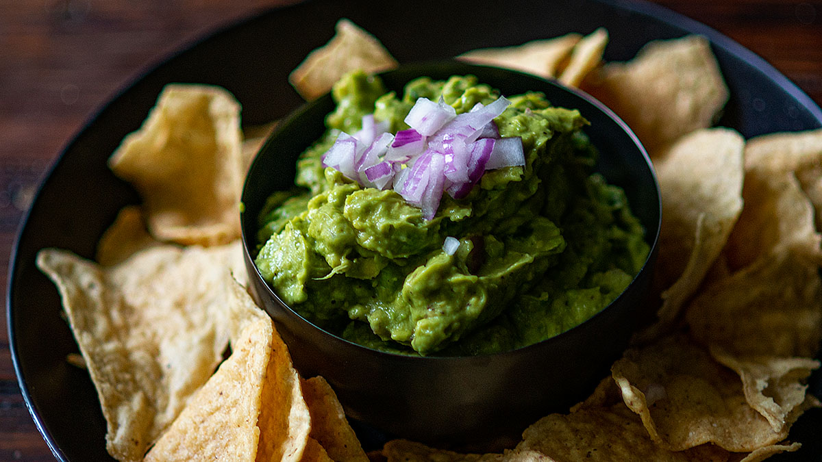 A bowl of guacamole topped with diced red onion, surrounded by tortilla chips on a dark plate.