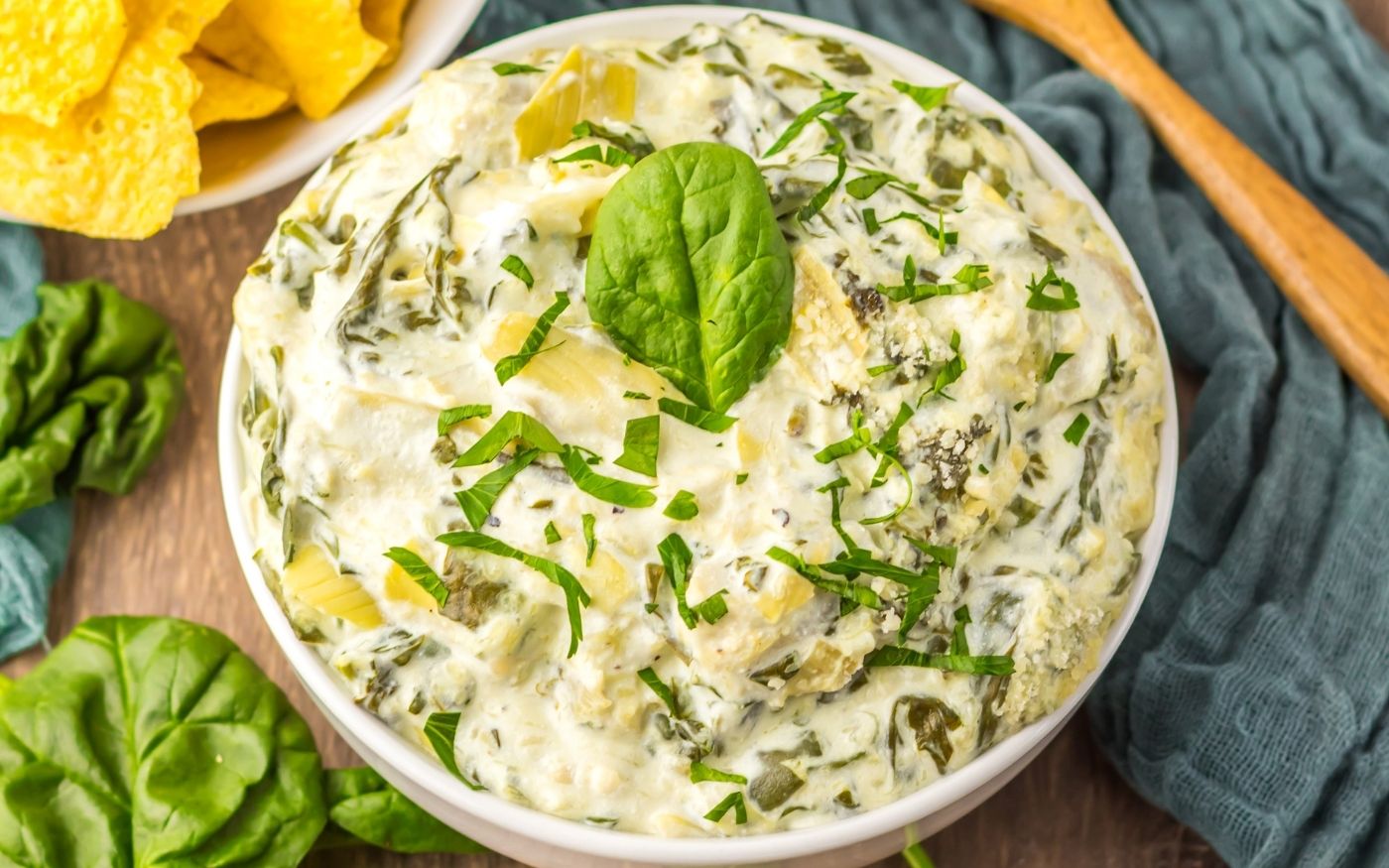 A bowl of creamy spinach artichoke dip garnished with chopped herbs and a fresh spinach leaf, with tortilla chips and spinach leaves nearby.