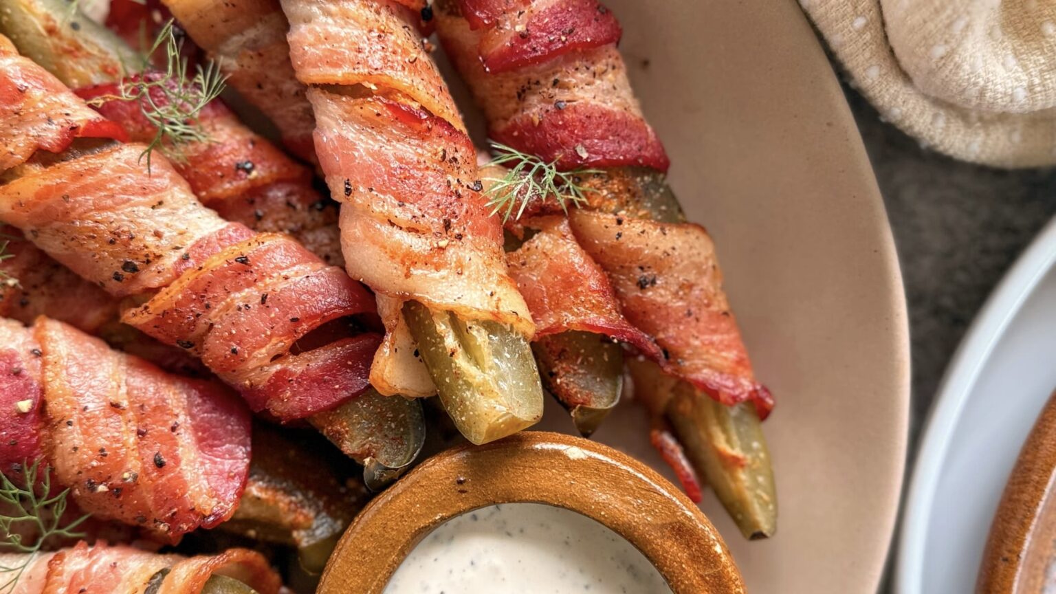 Bacon-wrapped pickles arranged on a plate next to a small bowl of creamy dipping sauce.