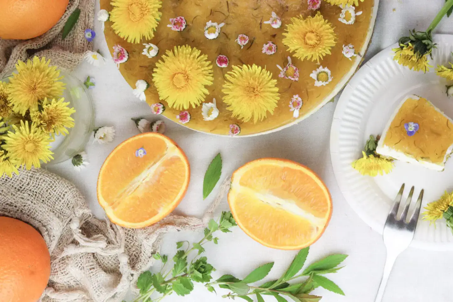 A cake topped with yellow flowers and small white blossoms, accompanied by orange halves, green leaves, and a slice of cake on a white plate with a fork.