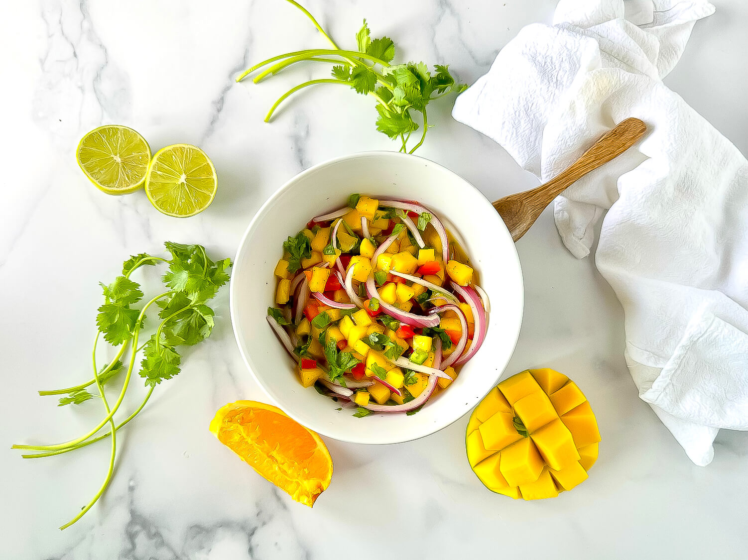 A bowl of mango salsa with red onion, cilantro, and diced pepper, surrounded by fresh cilantro, lime halves, a whole mango, and a white napkin on a marble surface.