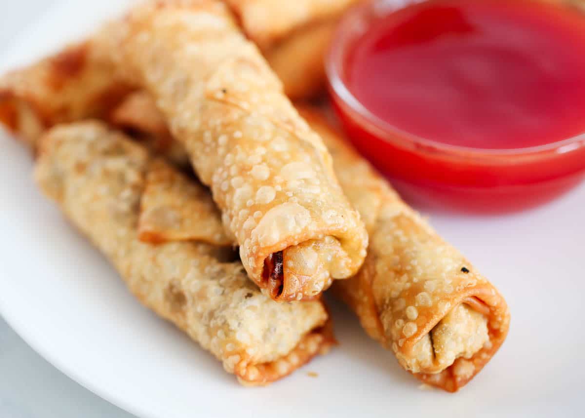 Three fried egg rolls are stacked on a white plate next to a small dish of red dipping sauce.