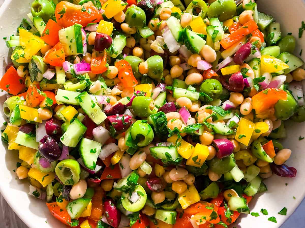 A close-up of a colorful chopped salad with cucumbers, olives, chickpeas, red onions, bell peppers, and parsley in a white bowl.