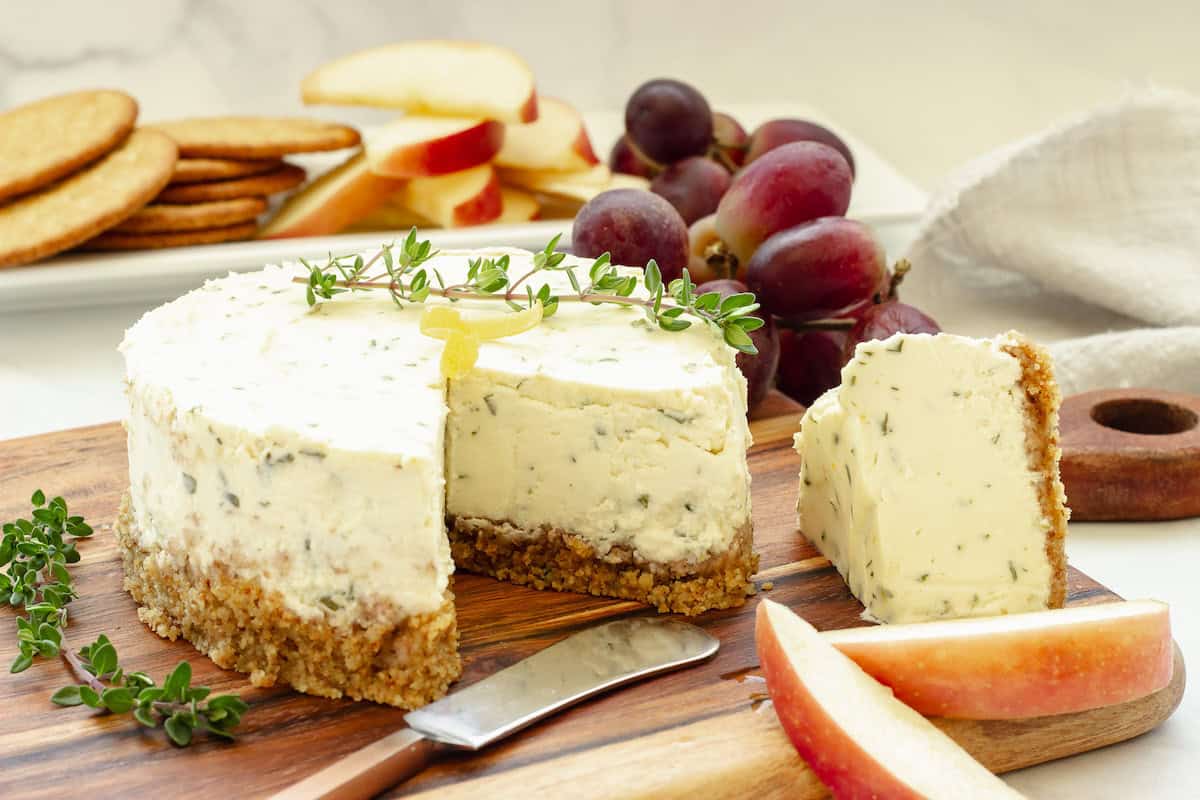 A round herbed cheese spread with a crumb crust sits on a wooden board, garnished with greens, with a slice cut out. Apple slices, grapes, and crackers are in the background.