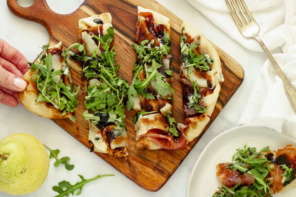 Flatbread topped with prosciutto, arugula, and balsamic glaze, sliced on a wooden board, with a hand taking a piece. A pear, fork, and plate with more flatbread are nearby.