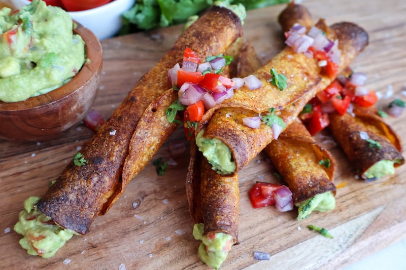 Three crispy rolled tacos filled with guacamole, garnished with diced tomatoes, onions, and herbs, served on a wooden board with a side of guacamole.