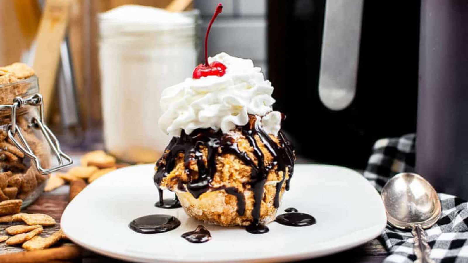 A dessert ball topped with whipped cream and a cherry, drizzled with chocolate sauce, sits on a white plate with a spoon and snacks in the background.