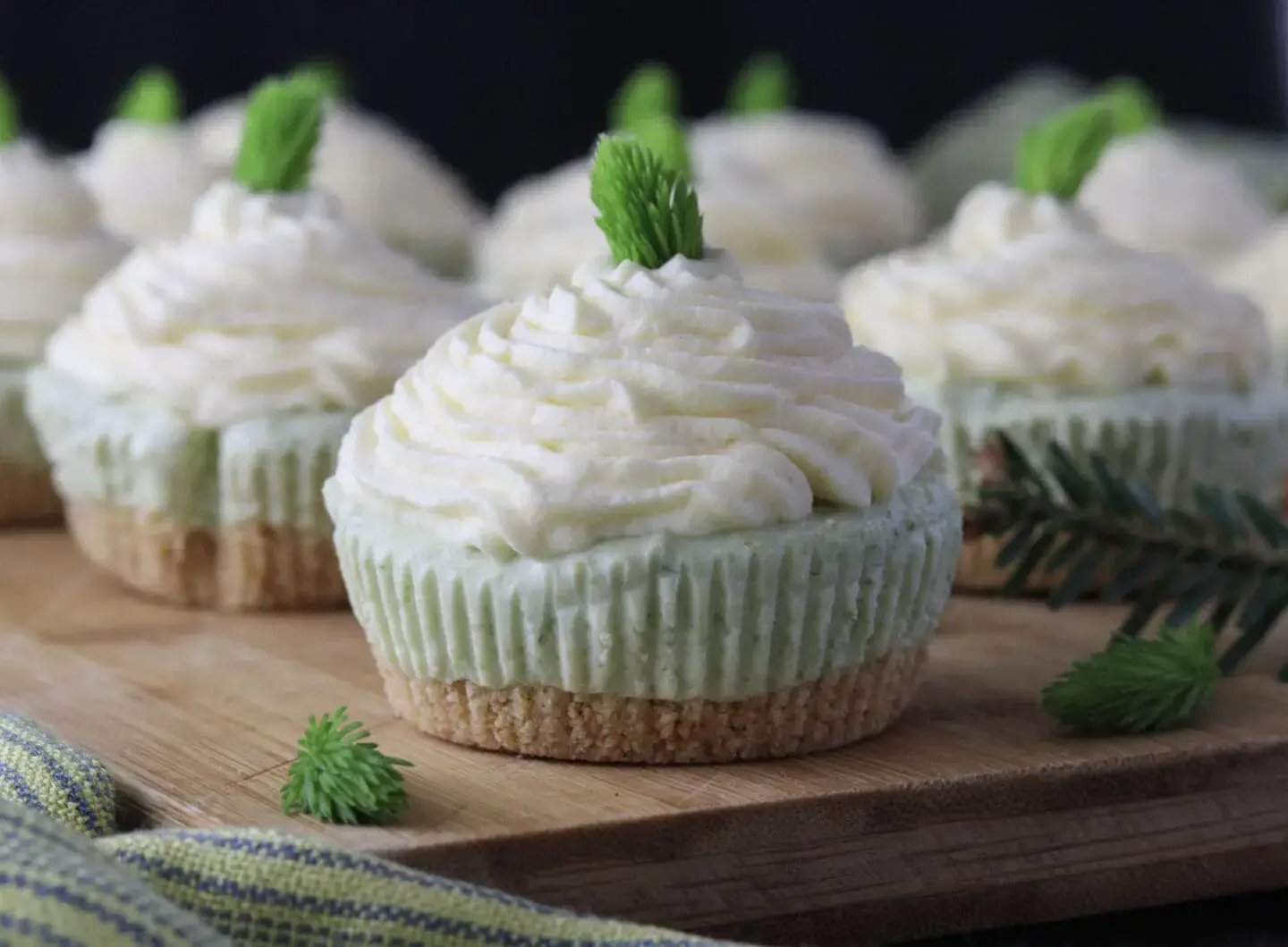 Mini cheesecakes with a green base and piped white topping, each garnished with a small green sprig, displayed on a wooden board.