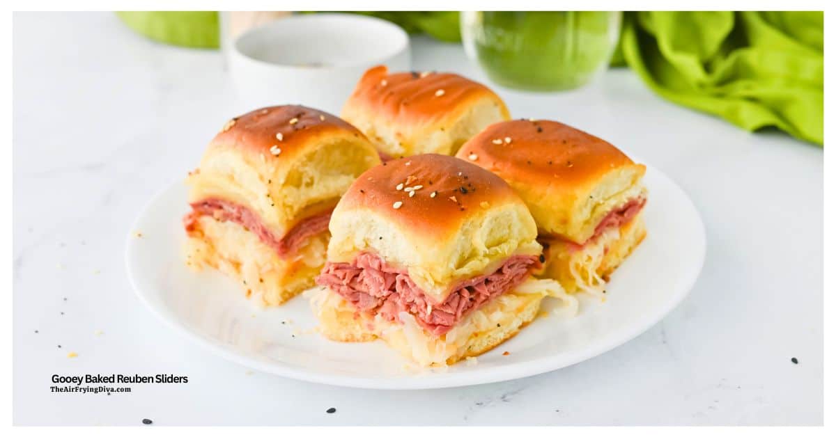 Four baked Reuben sliders with corned beef, sauerkraut, and melted cheese on soft rolls are arranged on a white plate.