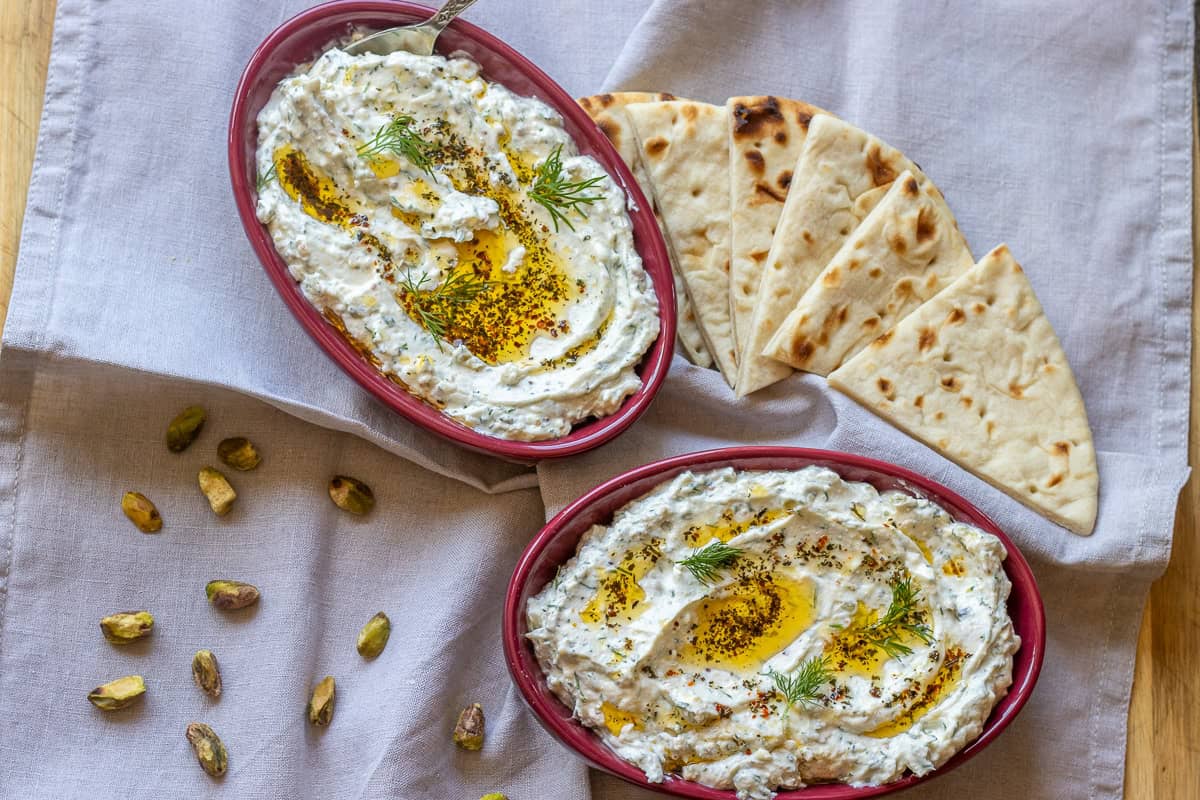 Two oval dishes of yogurt dip garnished with herbs and olive oil, served with sliced pita bread and scattered pistachios on a light cloth.