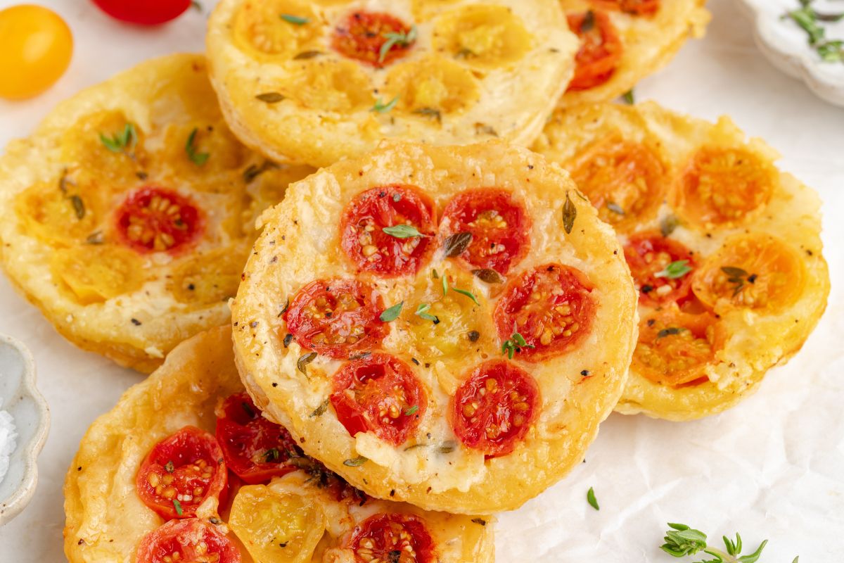 Mini tomato tarts with red and yellow cherry tomatoes, fresh herbs, and a golden pastry crust arranged on a white surface.