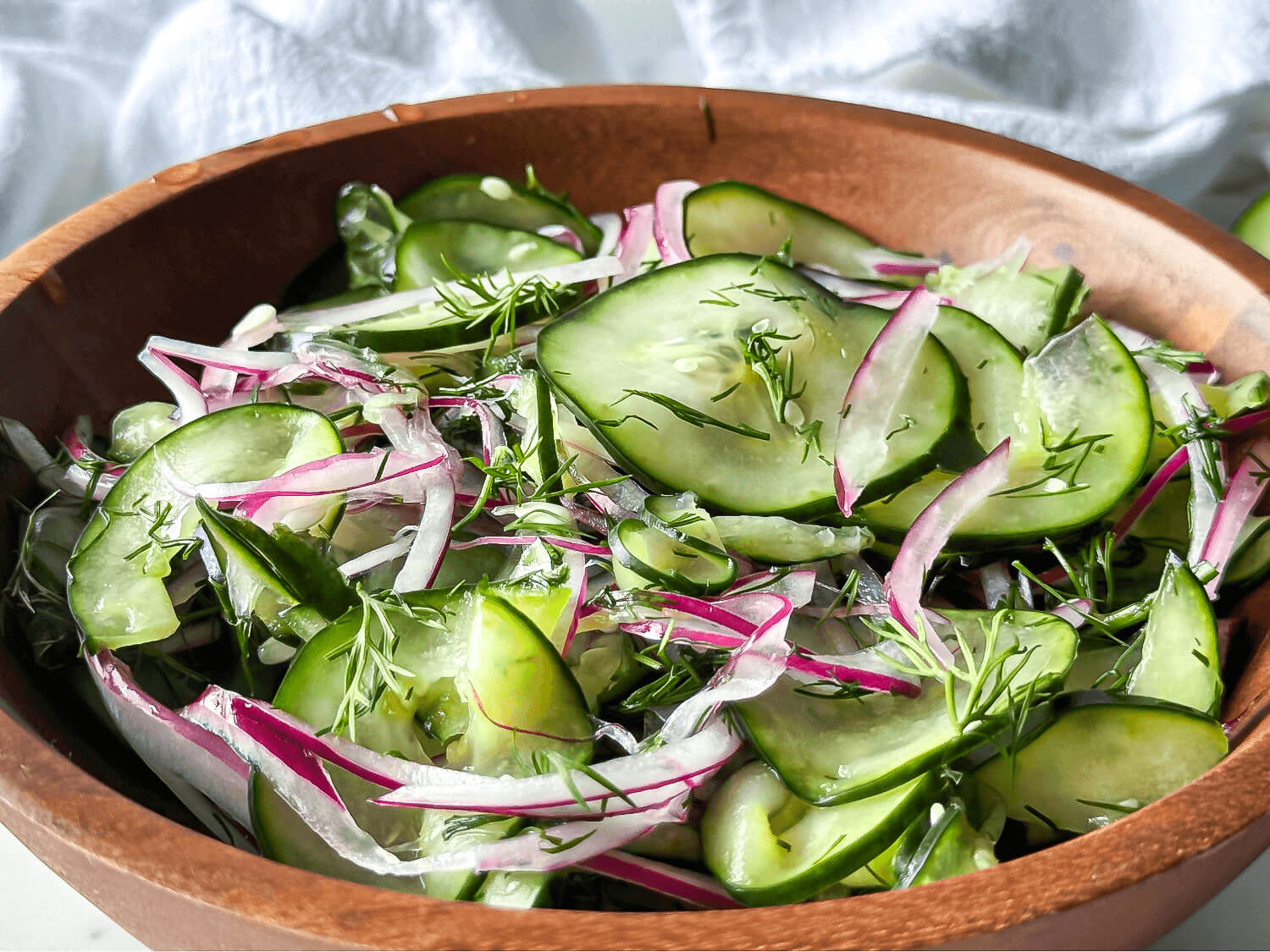 A wooden bowl filled with a salad of sliced cucumbers, red onions, and dill, mixed with a light dressing.