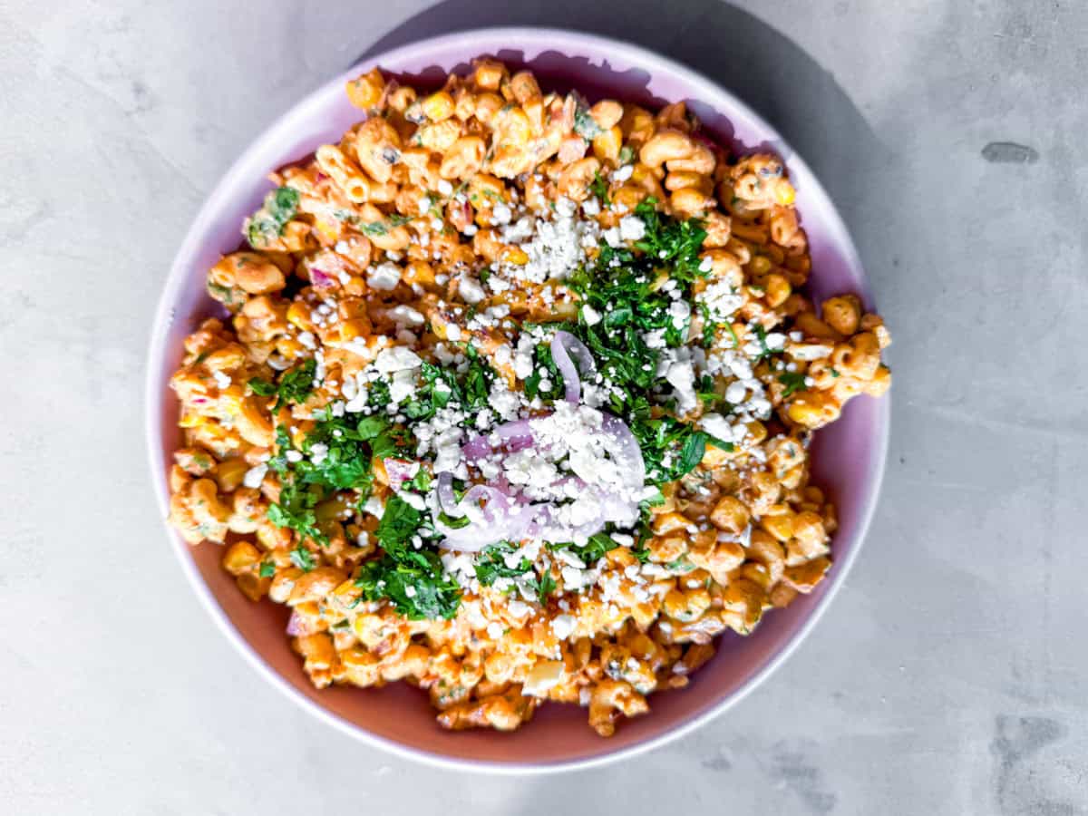 A bowl of creamy pasta salad topped with crumbled cheese, chopped herbs, and sliced red onions on a light gray surface.