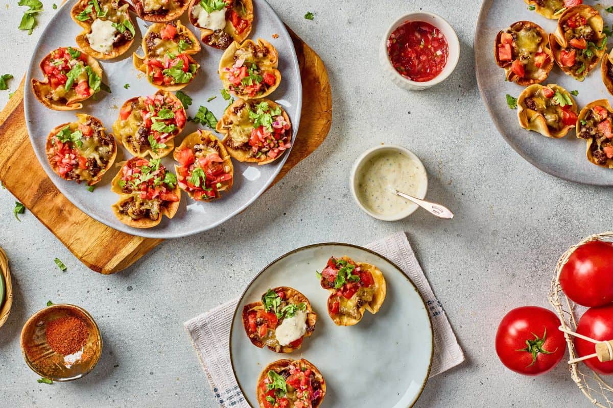 Several plates of mini taco cups topped with diced tomatoes, melted cheese, and herbs are arranged on a table with dipping sauces and fresh tomatoes.
