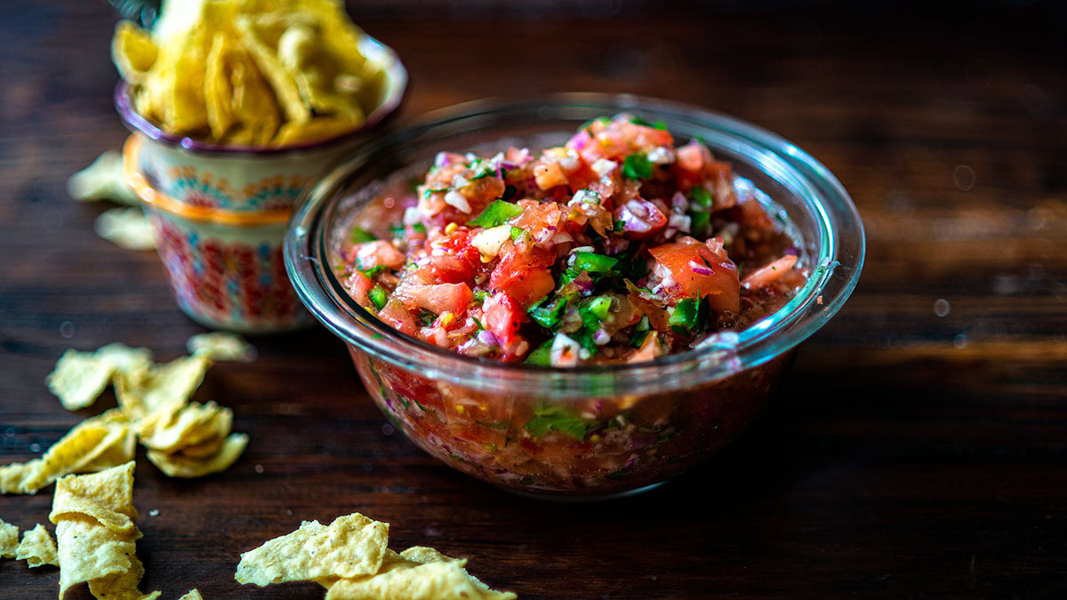 A clear glass bowl filled with freshly made salsa sits on a wooden table, with tortilla chips in a ceramic container and scattered nearby.
