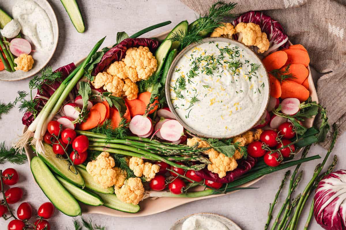 A platter of assorted fresh vegetables including carrots, cauliflower, asparagus, radishes, cucumbers, and cherry tomatoes with a bowl of creamy dip garnished with herbs.
