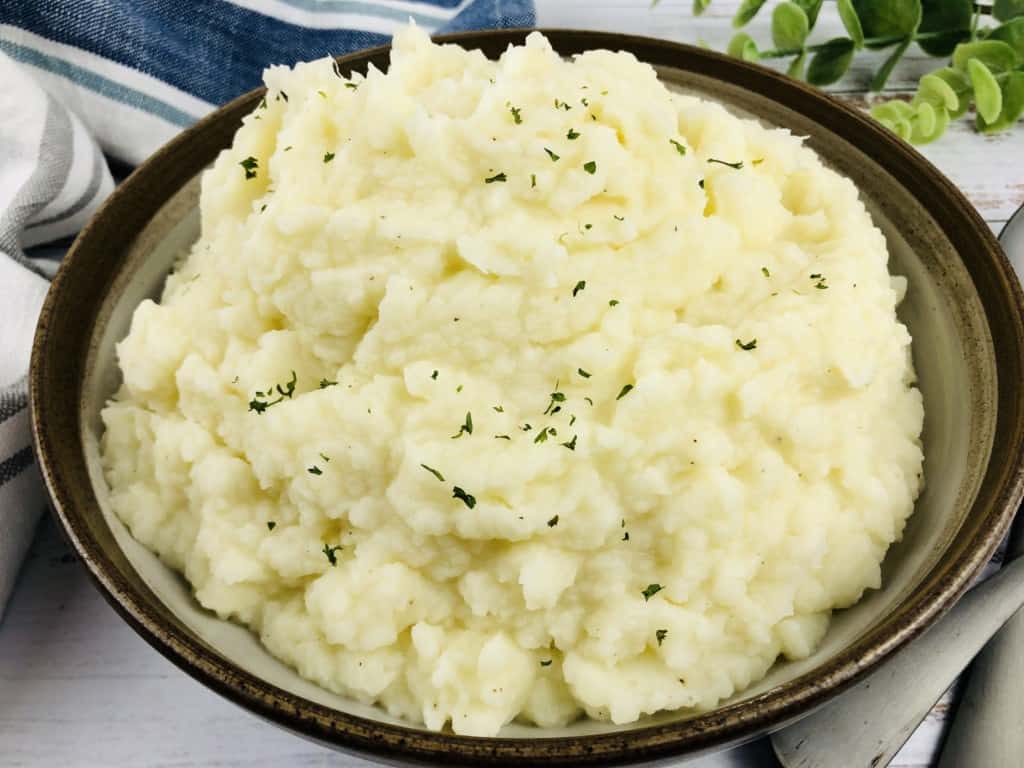 A bowl of creamy mashed potatoes garnished with finely chopped herbs, placed on a white surface with a striped cloth and green plant nearby.