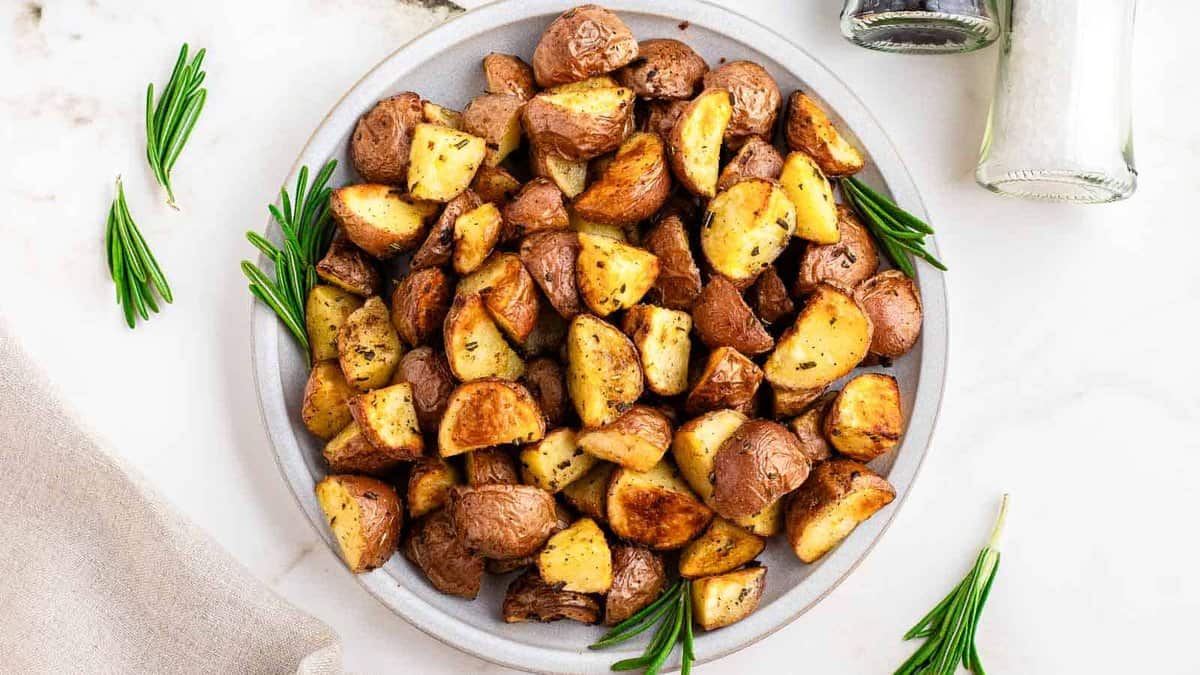 A plate of roasted potatoes with herbs, surrounded by sprigs of rosemary and salt and pepper shakers on a white surface.