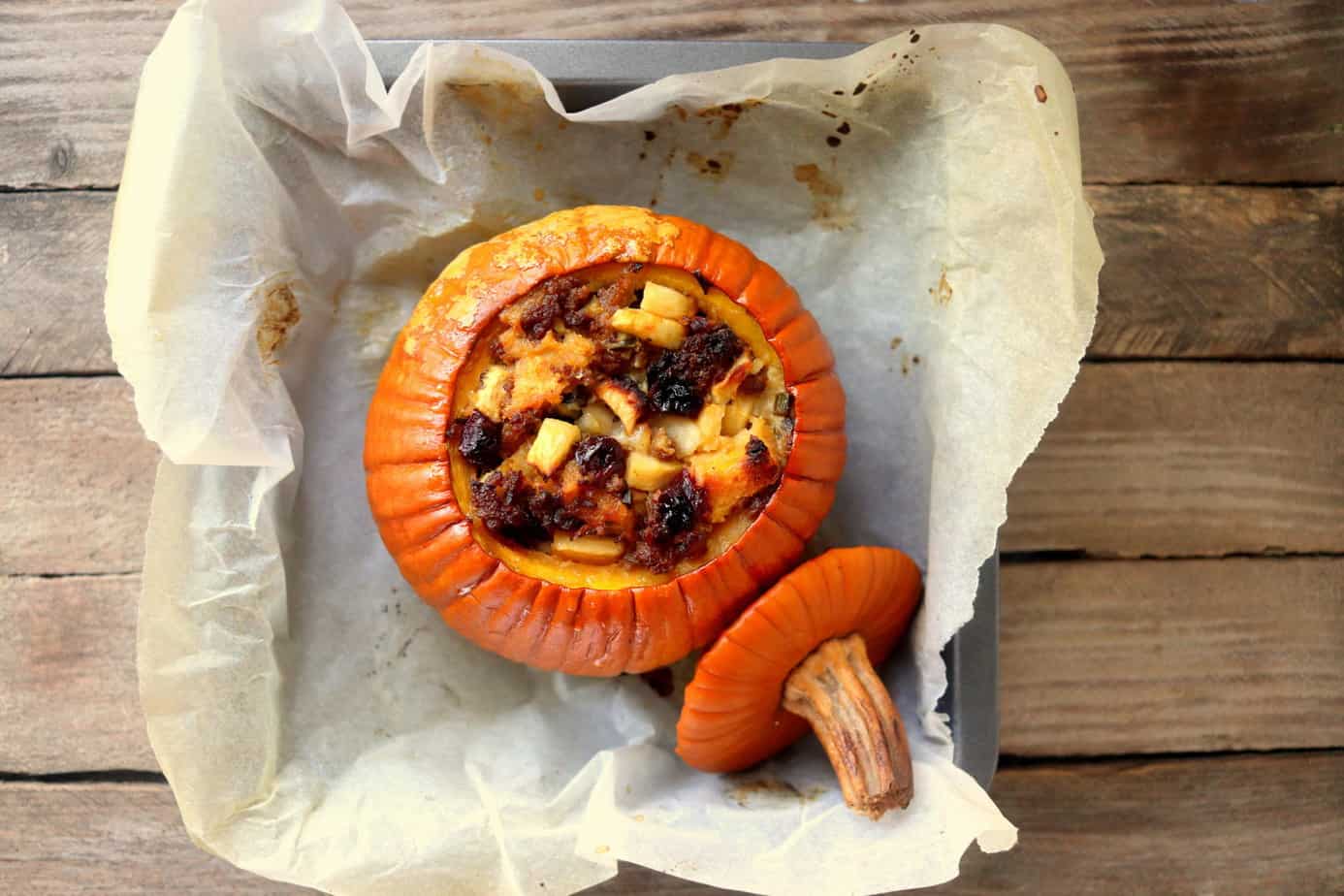 A whole pumpkin with its top removed and set aside, filled with a baked savory mixture, sits on parchment paper in a square baking dish on a wooden surface.