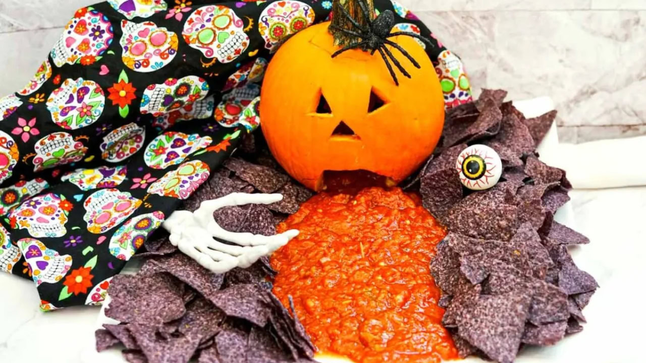 A carved pumpkin with a distressed expression, placed among blue corn chips, appears to vomit salsa; a fake skeleton hand and eyeball are also present.