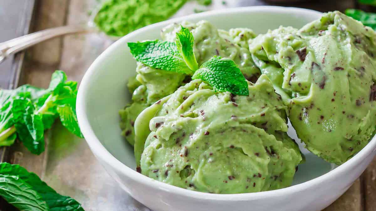 Three scoops of mint chocolate chip ice cream in a white bowl, garnished with fresh mint leaves.