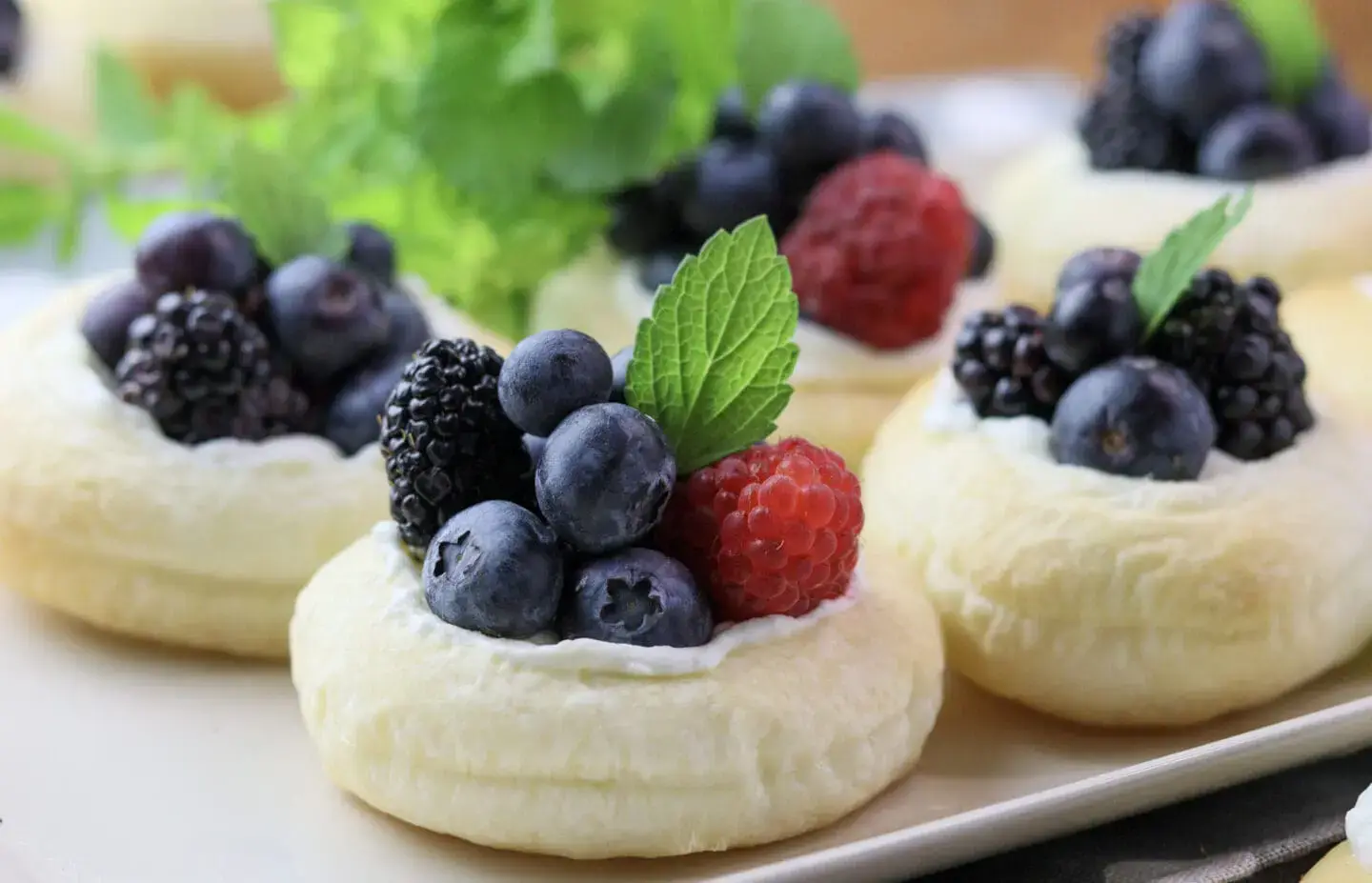 Mini puff pastry tarts topped with cream, assorted berries, and mint leaves are arranged on a white plate.