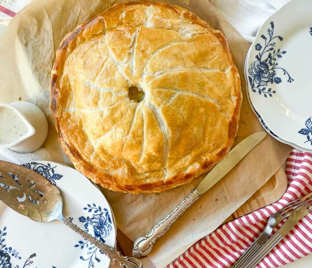A golden-brown pie with a flaky crust sits on parchment paper next to a floral plate, serving utensils, and a red-striped napkin.