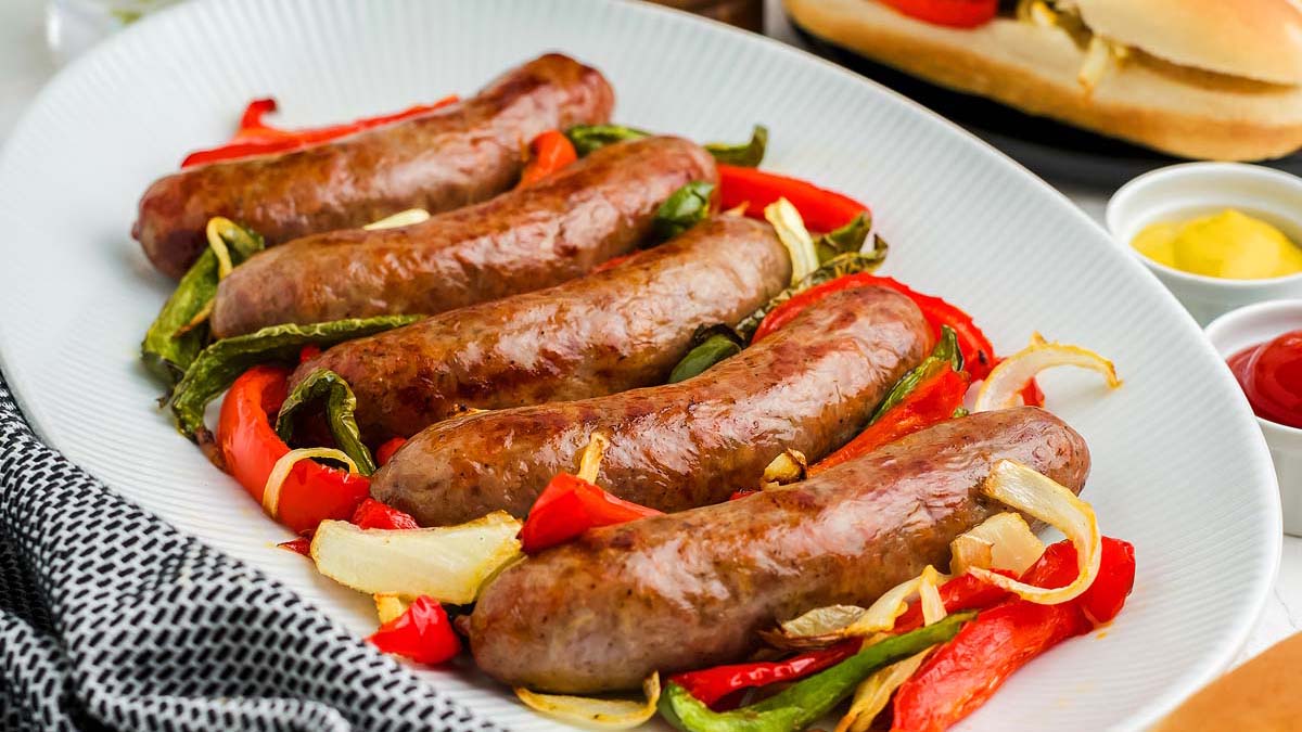 Five grilled sausages served on a white plate with sliced red and green bell peppers and onions.