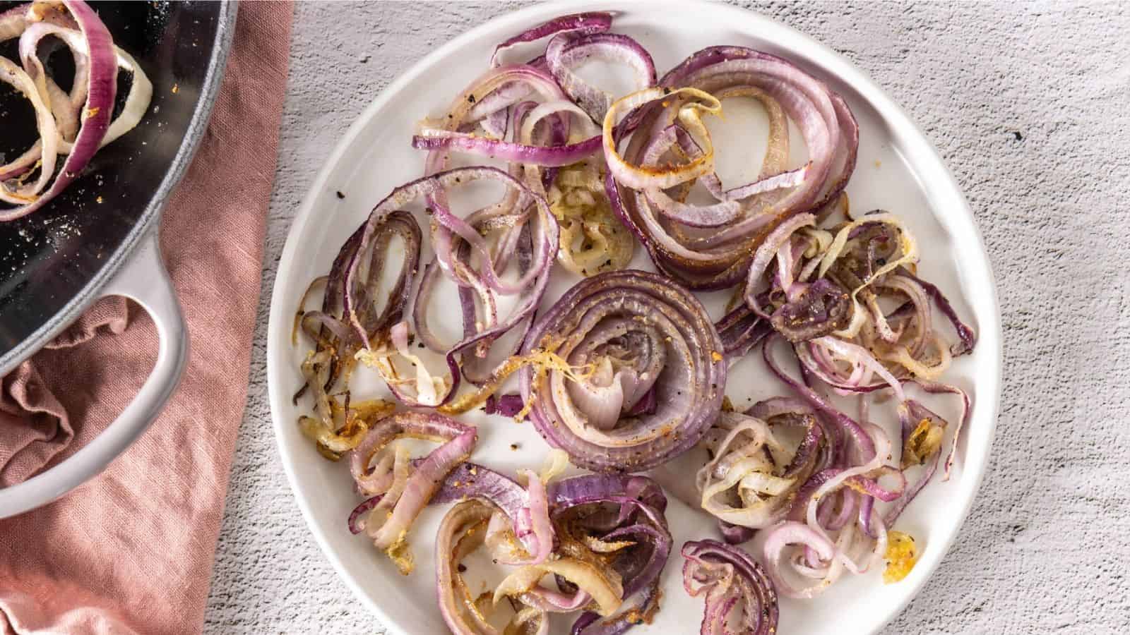 A white plate with cooked, sliced red onions, some slightly caramelized, on a light textured surface next to a pink cloth and a skillet.