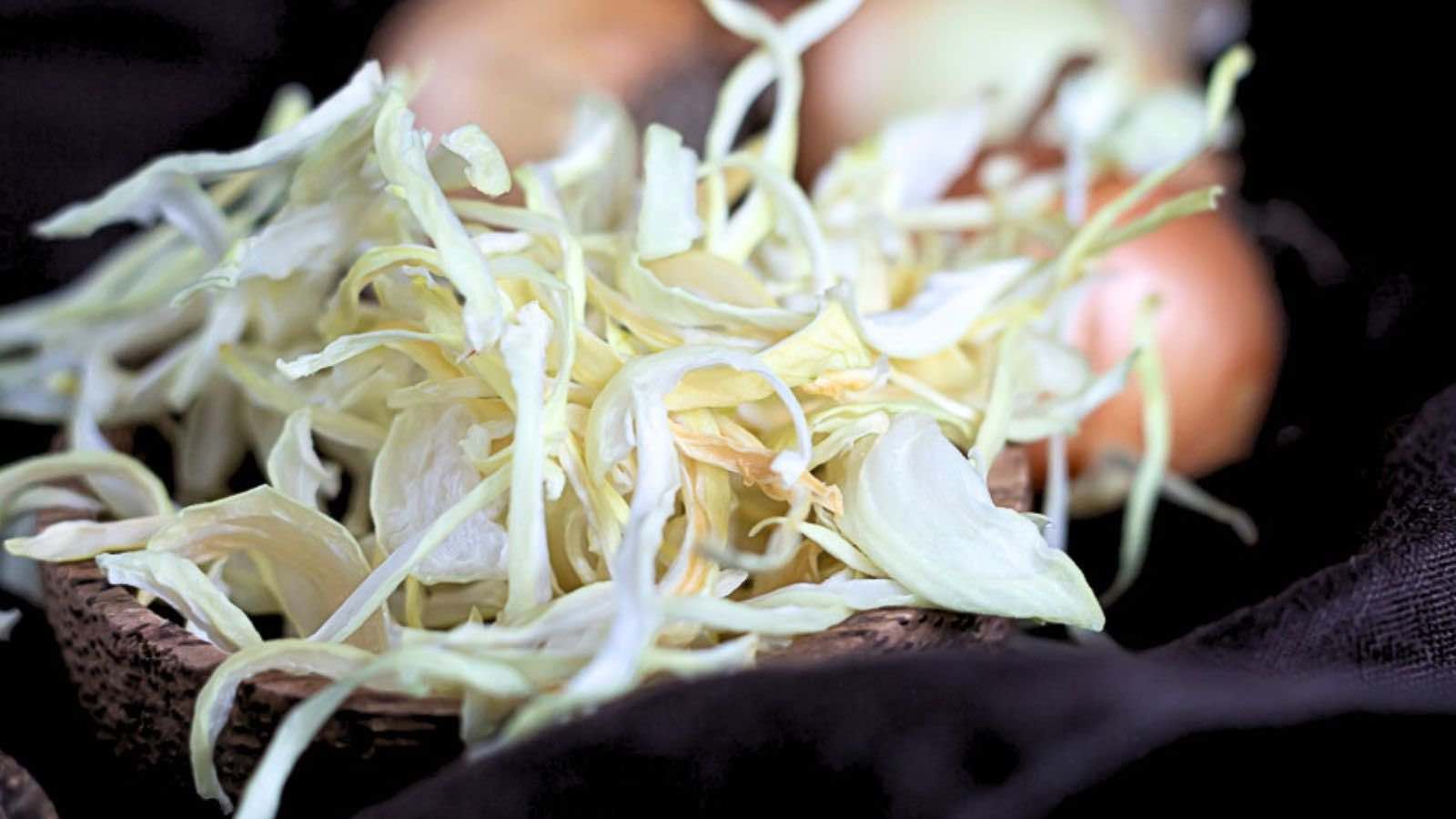 A close-up of a wooden bowl filled with shredded cabbage, with some whole onions blurred in the background.