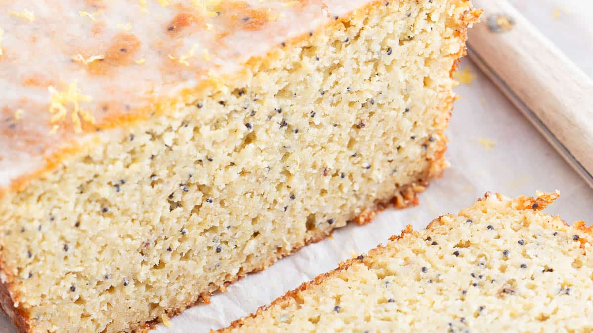 A close-up of a sliced loaf of lemon poppy seed cake on parchment paper, showing a moist, speckled crumb.