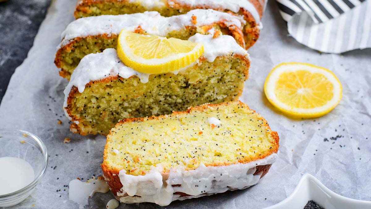 Sliced lemon poppy seed loaf with white glaze, topped with lemon slices, displayed on parchment paper with a lemon slice and glaze nearby.