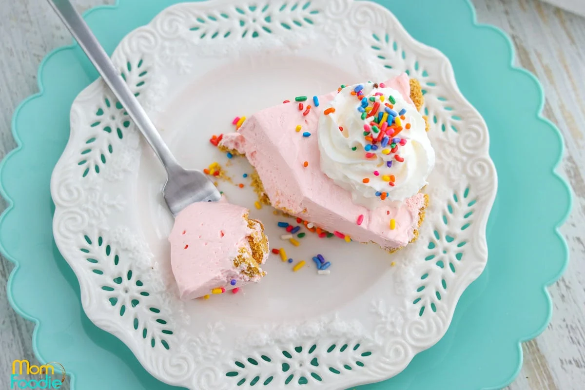 A slice of pink pie with whipped cream and rainbow sprinkles on a white plate, with a fork holding a bite-sized piece.