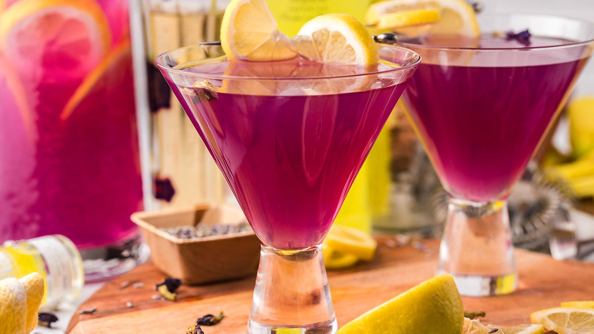 Two martini glasses filled with bright purple drinks, each garnished with lemon slices, sit on a wooden surface with lemons and a pitcher in the background.