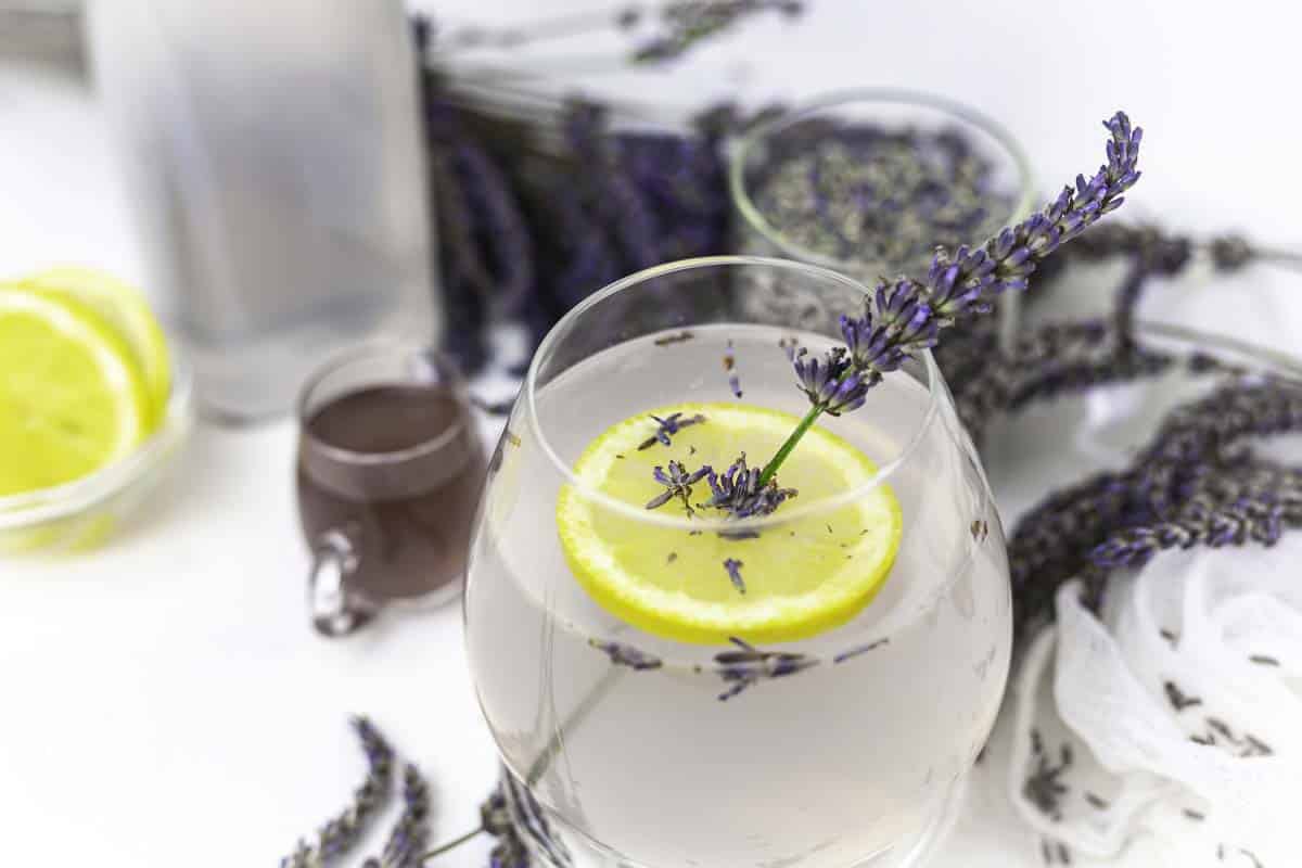 A glass of lavender lemonade garnished with lemon slices and a sprig of lavender, surrounded by lavender flowers, a cup of syrup, and lemon slices.