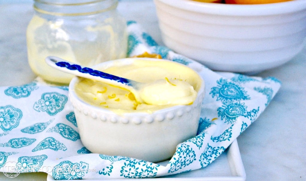 A small white bowl filled with creamy yellow lemon curd, garnished with lemon zest, sits on a patterned napkin with a spoon inside. A jar and a bowl of fruit are in the background.