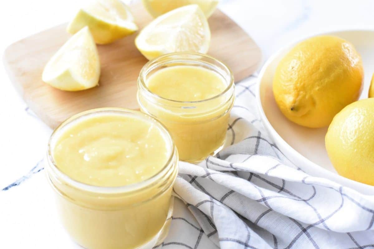Two jars of lemon curd sit on a cloth next to whole and sliced lemons on a cutting board.