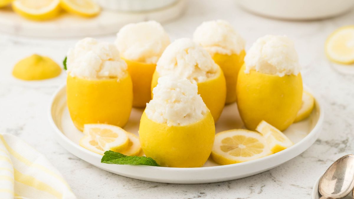 Five hollowed-out lemons filled with lemon sorbet are arranged on a white plate, garnished with fresh lemon slices and a mint leaf.