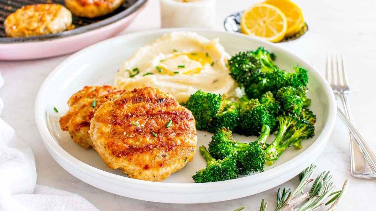 A white plate with two chicken patties, mashed potatoes topped with olive oil, and roasted broccoli, with a fork and sliced lemons in the background.