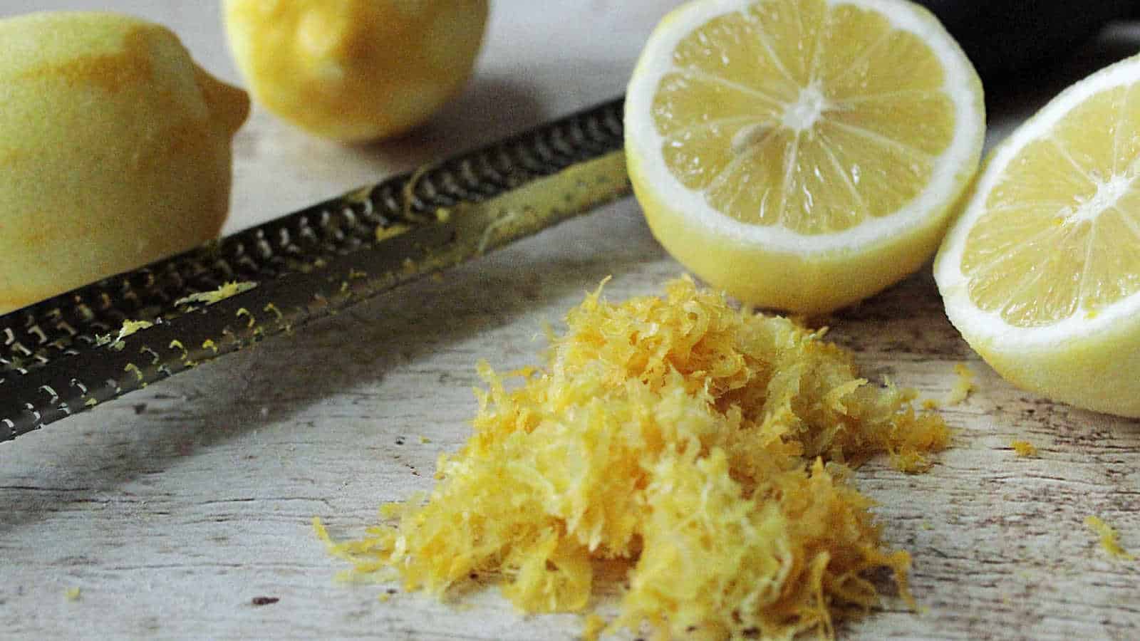 Whole lemons, a zester with lemon zest, and two lemon halves are arranged on a wooden surface.