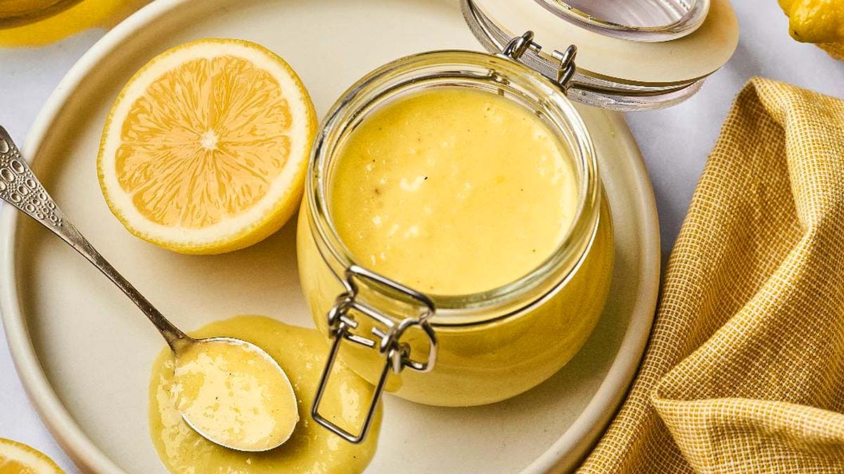 An open jar of lemon curd on a plate with a halved lemon, a spoon with curd, and a yellow checked cloth nearby.