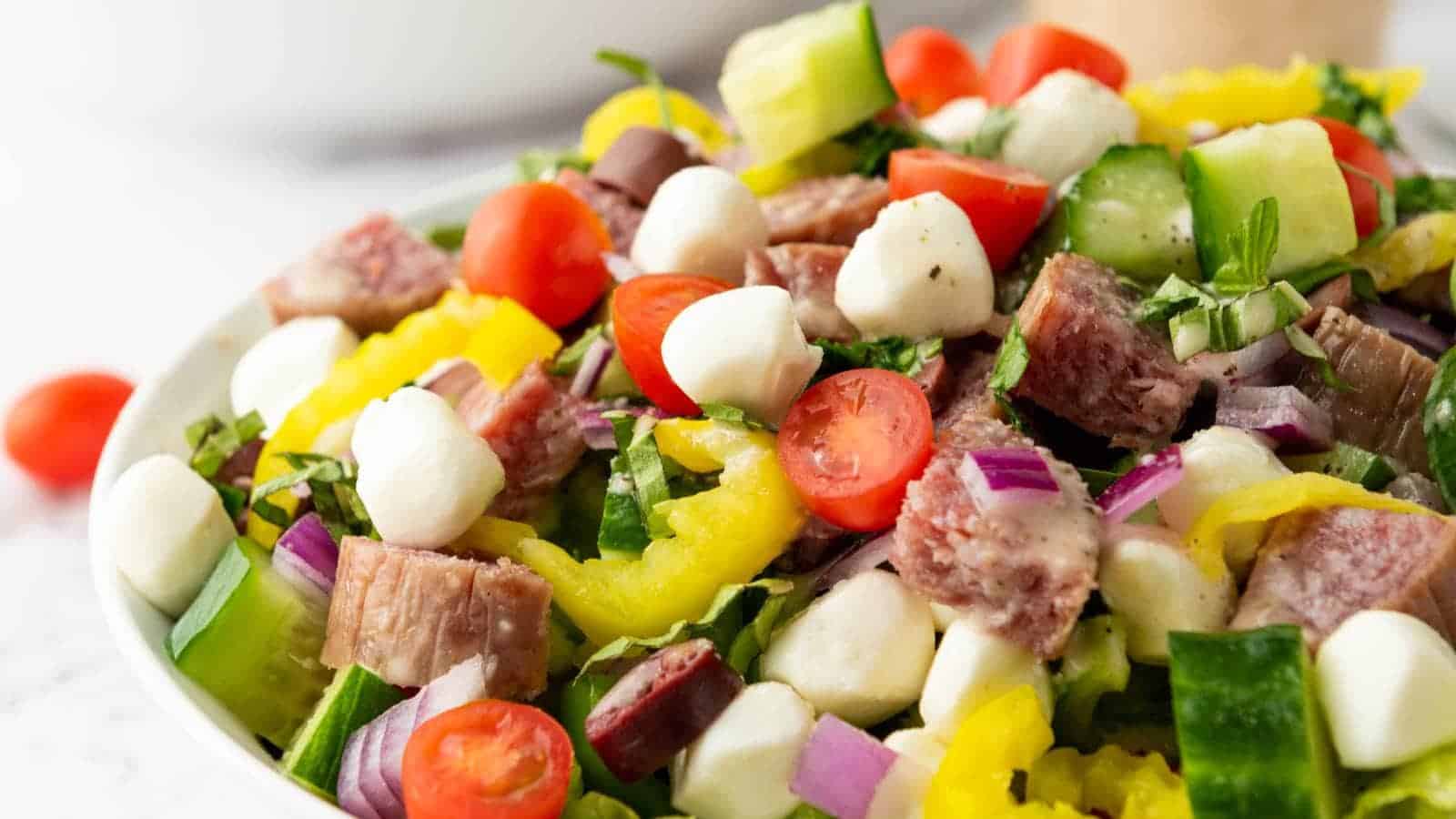 A bowl of salad with cherry tomatoes, mozzarella balls, cucumber, yellow peppers, red onion, leafy greens, and chunks of cured meat.