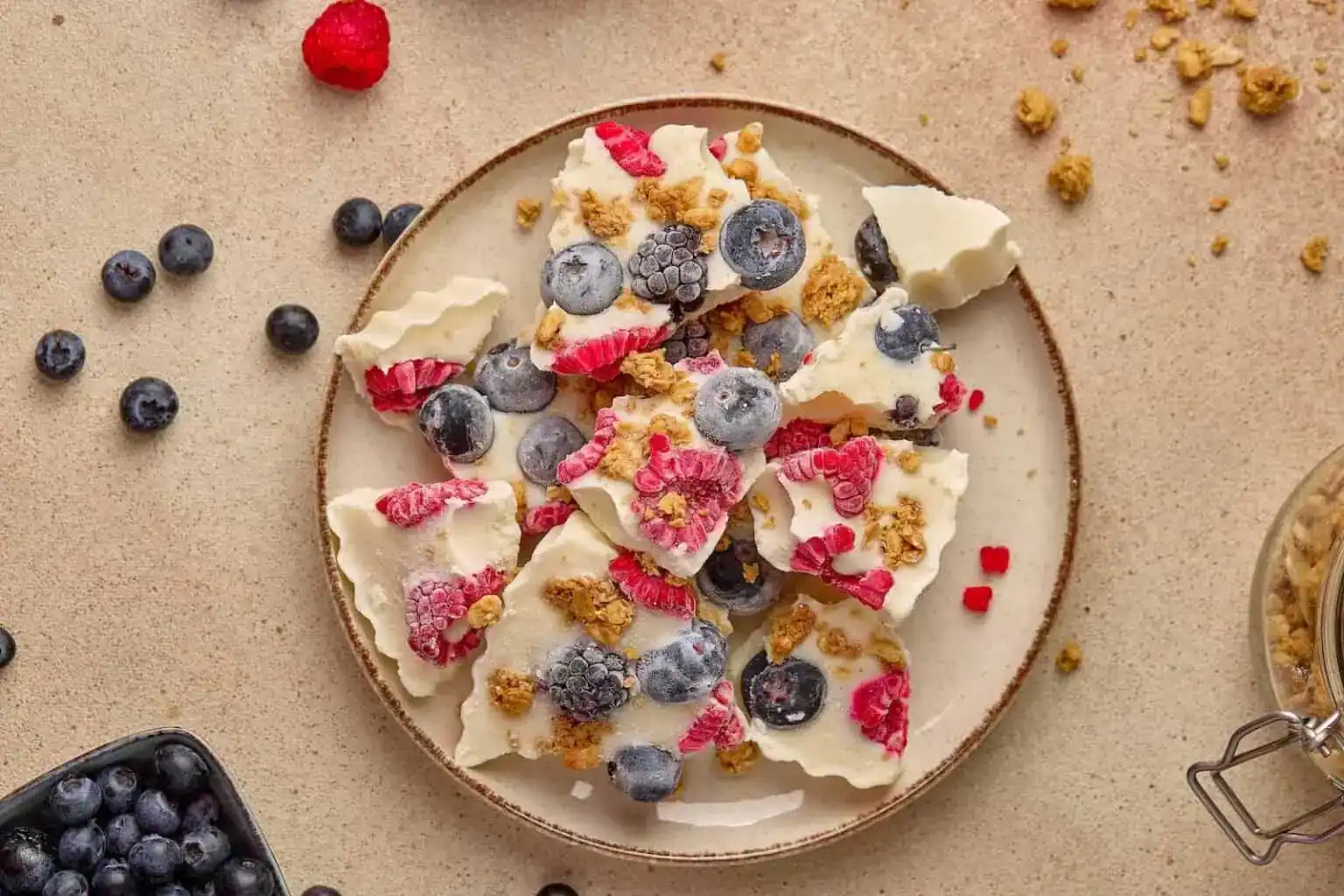 A plate of yogurt bark topped with blueberries, raspberries, and granola pieces, arranged on a light-colored surface.