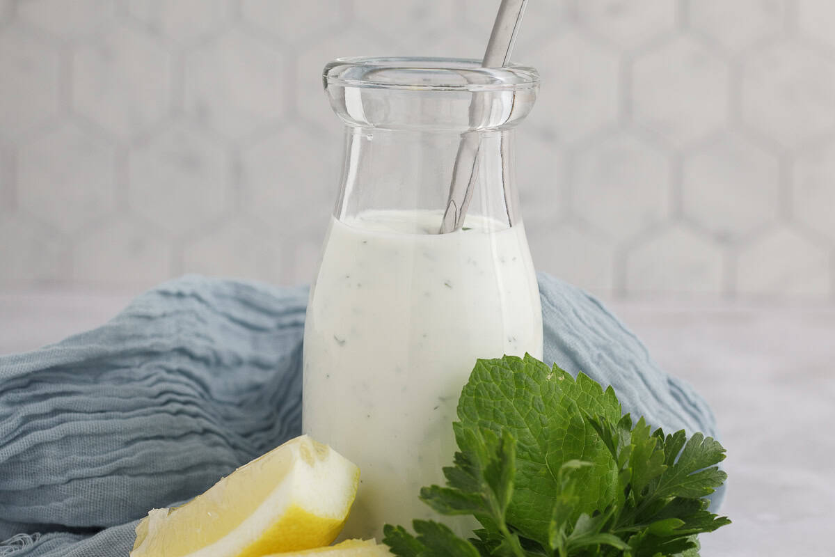 A glass bottle filled with creamy white dressing, a metal spoon inside, placed next to lemon wedges and fresh green herbs on a blue cloth.
